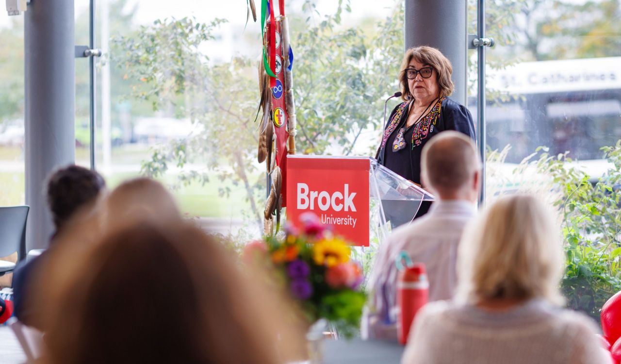 A woman stands in front of a Brock University-branded lectern with a wall of windows behind her. She is speaking into a microphone, addressing a crowd of people seated at tables.