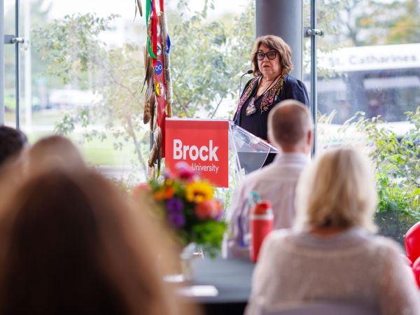 A woman stands in front of a Brock University-branded lectern with a wall of windows behind her. She is speaking into a microphone, addressing a crowd of people seated at tables.