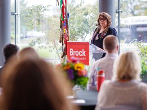 A speaker stands at a Brock University podium addressing an audience during an indoor event. Large windows, colourful flowers and an Indigenous ceremonial display are visible in the background.