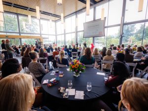 Audience members seated at round tables listen to speakers during a formal event in a bright, glass-walled atrium. A presentation screen is displayed at the front of the room, with floral centrepieces and red-and-white balloons visible among the attendees.