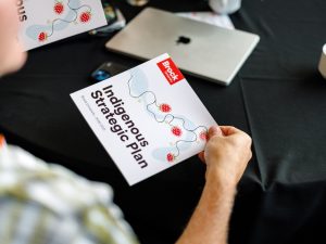 A person seated at a table holds a copy of Brock University’s Indigenous Strategic Plan 2026–2031. The booklet cover features strawberry illustrations and flowing line designs, with a laptop, phone and a second copy of the plan visible on the table in the background.