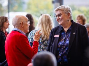 Two people speak at a campus gathering inside a bright, window-lined space. One person in a red sweater gestures while talking with another person wearing glasses, a dark blazer and a Brock name tag as attendees mingle in the background.