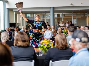 A dancer wearing brightly coloured regalia performs in the centre of a crowd gathered in an indoor event space, holding a feather fan overhead while attendees seated at round tables watch.
