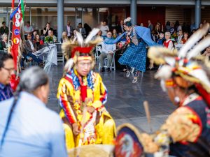 A dancer in blue regalia performs in the centre of a large indoor gathering space while drummers and singers seated in the foreground play a large drum. Audience members watch from tables and chairs surrounding the performance area, and Indigenous flags stand nearby.