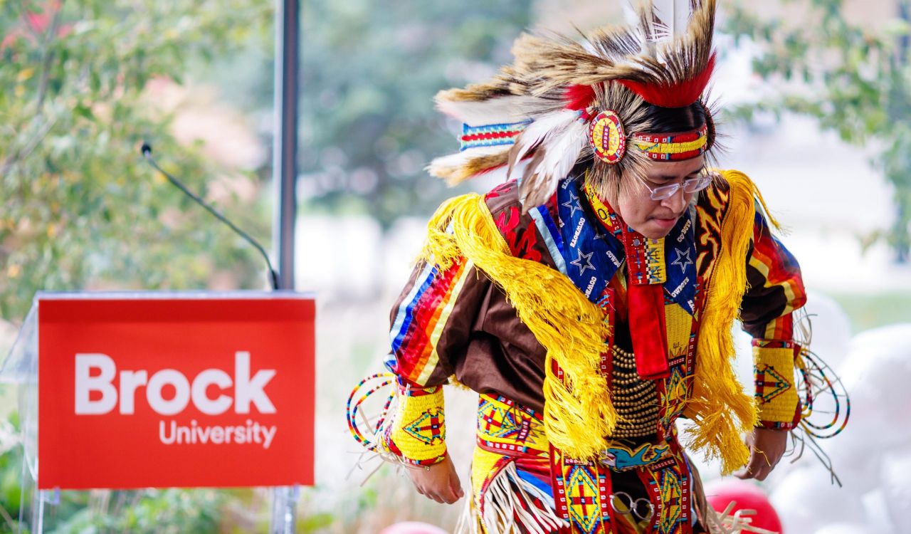 A dancer wearing brightly coloured regalia with beadwork, fringe and a feathered headdress performs near a Brock University podium during an indoor event. Red and white balloons are visible in the background.