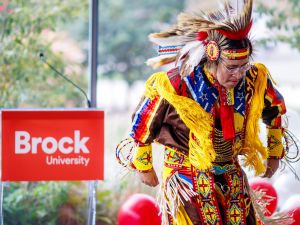 A dancer wearing brightly coloured regalia with beadwork, fringe and a feathered headdress performs near a Brock University podium during an indoor event. Red and white balloons are visible in the background.