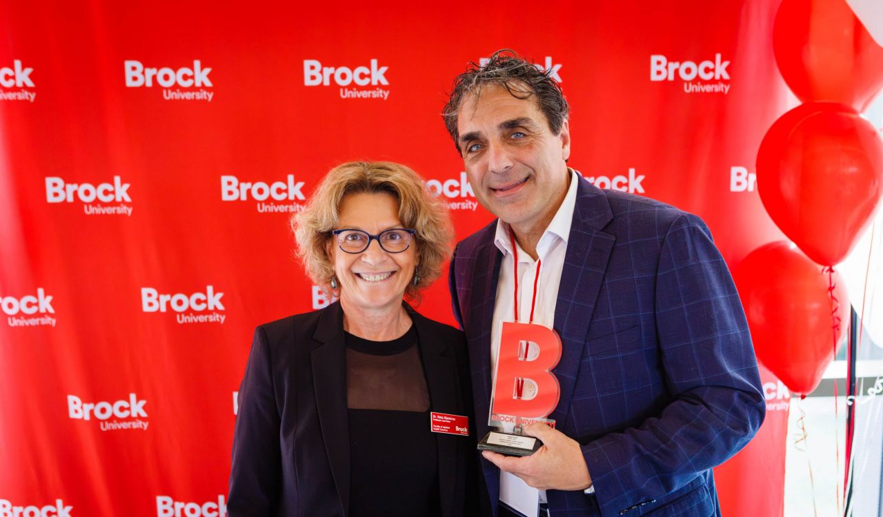 A woman and a man, holding a trophy, pose in front of a red backdrop.