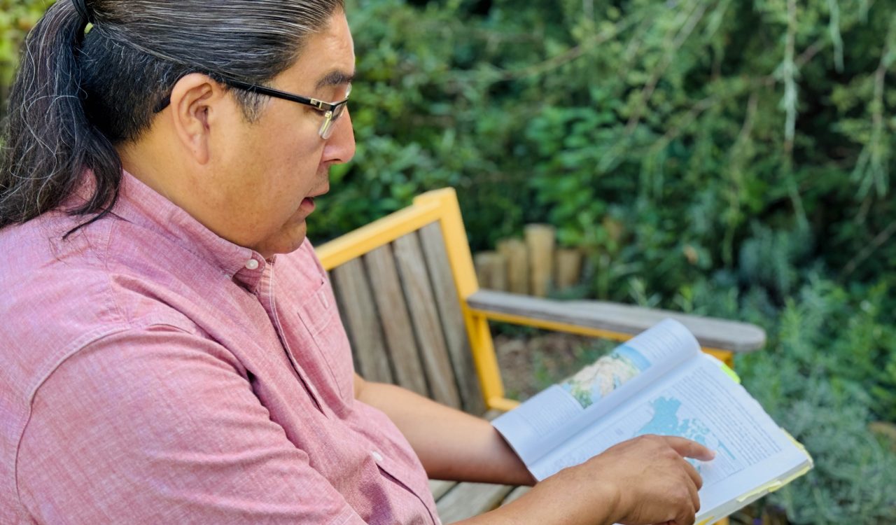 Joshua Manitowabi sits outside at Brock University reading a book containing a map.