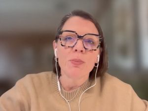 A screenshot from a CBC podcast video of a female guest sitting in an office, wearing glasses and headphones.