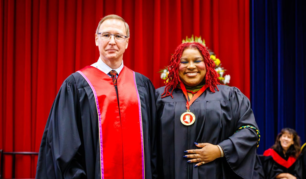 Two people in academic robes pose for a photo on stage during a university graduation ceremony.