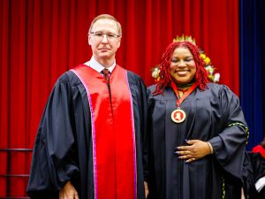 Two people in academic robes pose for a photo on stage during a university graduation ceremony.