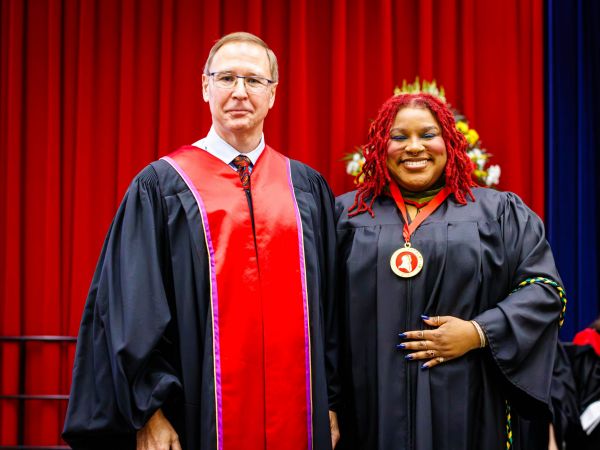 Two people in academic robes pose for a photo on stage during a university graduation ceremony.