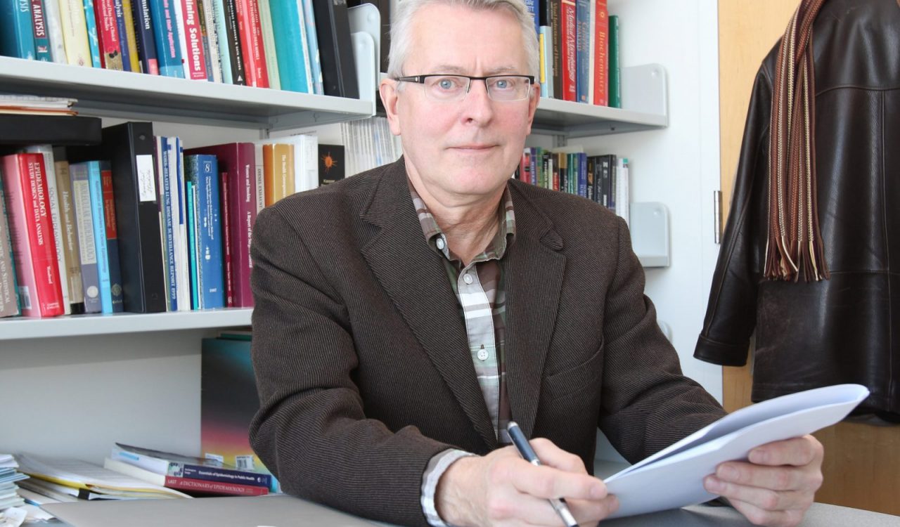 Martin Tammemägi sits at a desk holding a pen and a stack of papers, with a shelf of books and his coat hanging in the background.