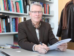 Martin Tammemägi sits at a desk holding a pen and a stack of papers, with a shelf of books and his coat hanging in the background.