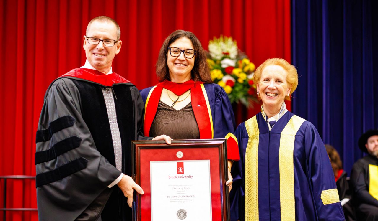 Three people in academic robes pose with a framed diploma on stage during a university graduation ceremony.