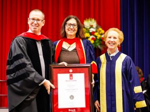 Three people in academic robes pose with a framed diploma on stage during a university graduation ceremony.