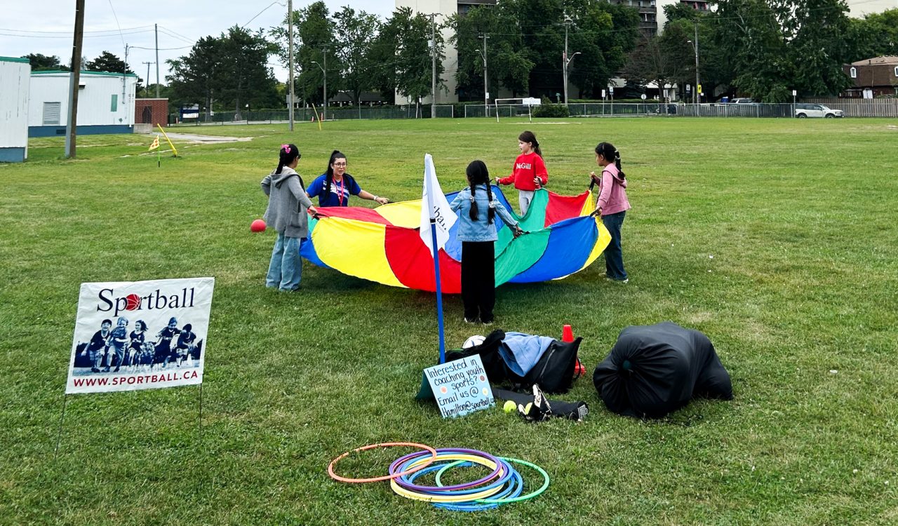 A circle of girls holds up a multi-coloured tarp in a green field, with hoola-hoops, other equipment and a sign saying "Sportball" in the foreground.