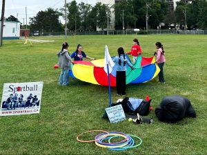A circle of girls holds up a multi-coloured tarp in a green field, with hoola-hoops, other equipment and a sign saying "Sportball" in the foreground.