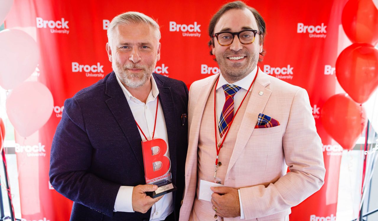 Two men in suits pose with an award in front of a red backdrop.