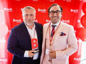 Two men in suits pose with an award in front of a red backdrop.