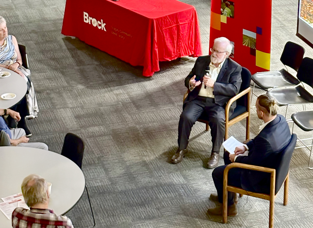 Two men sit inside the Pond Inlet in chairs angled slightly toward one another but facing out to an audience. One holds a mic and speaks to the audience, while the other looks on.