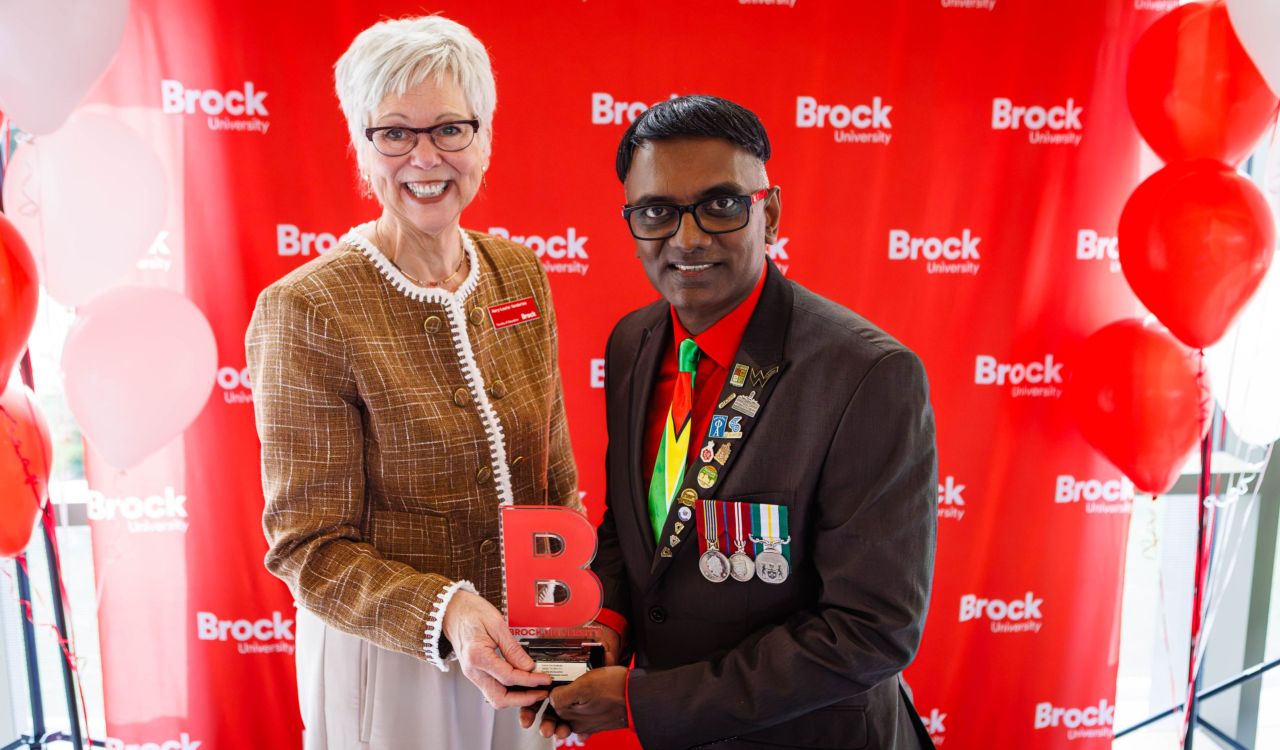 Mary-Louise Vanderlee hands Dr. Narine Dat Sookram a small award standing in front of a Brock University banner.