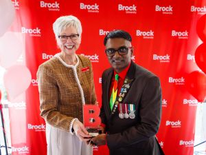Mary-Louise Vanderlee hands Dr. Narine Dat Sookram a small award standing in front of a Brock University banner.
