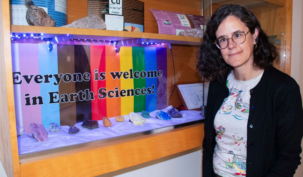 A woman stands beside a wall-mounted display cabinet with colourful minerals on display in front of a rainbow-print sign featuring the words “Everyone is welcome in Earth Sciences!” in bold lettering.