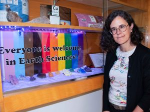 A woman stands beside a wall-mounted display cabinet with colourful minerals on display in front of a rainbow-print sign featuring the words “Everyone is welcome in Earth Sciences!” in bold lettering.