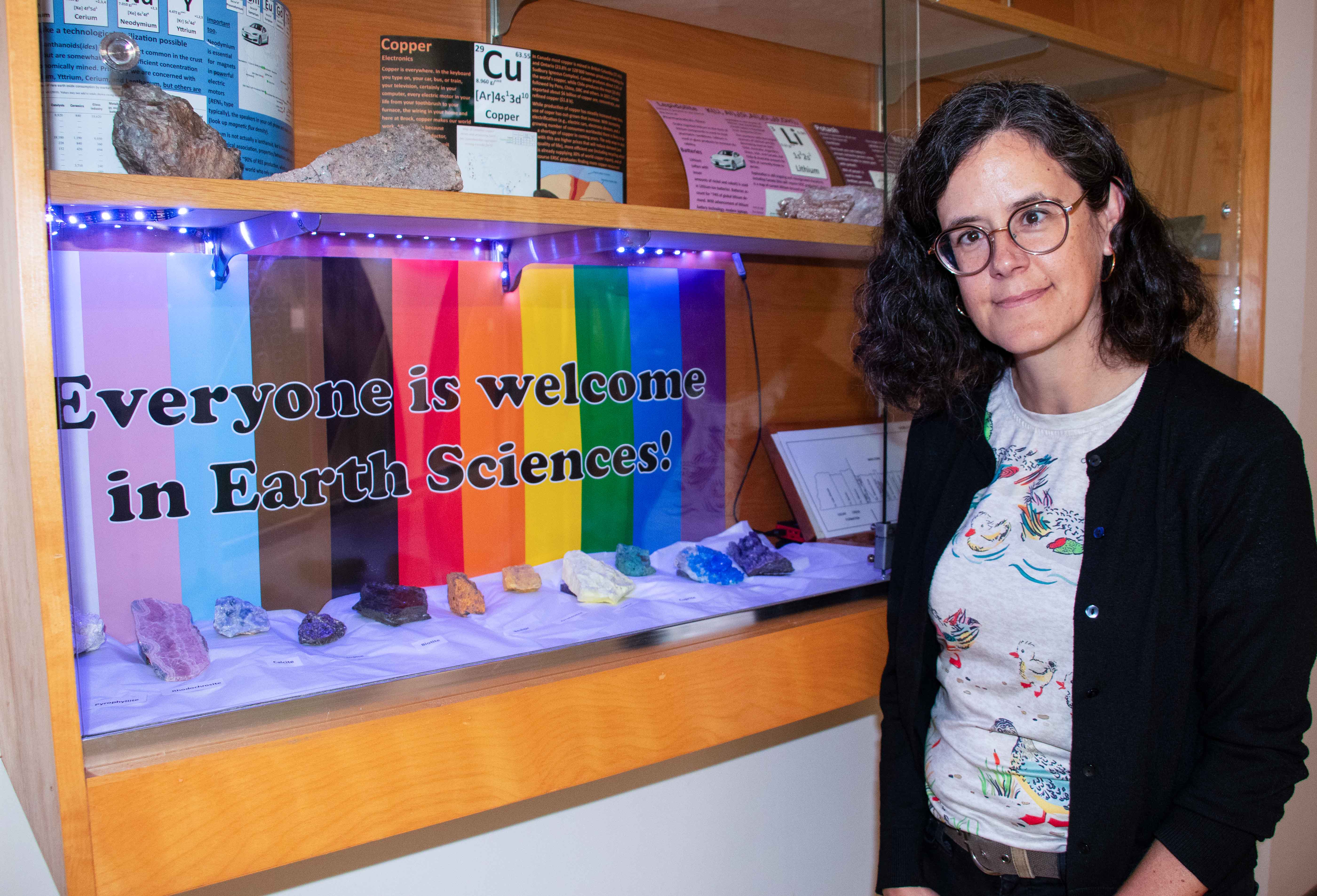 A woman stands beside a wall-mounted display cabinet with colourful minerals on display in front of a rainbow-print sign featuring the words “Everyone is welcome in Earth Sciences!” in bold lettering.