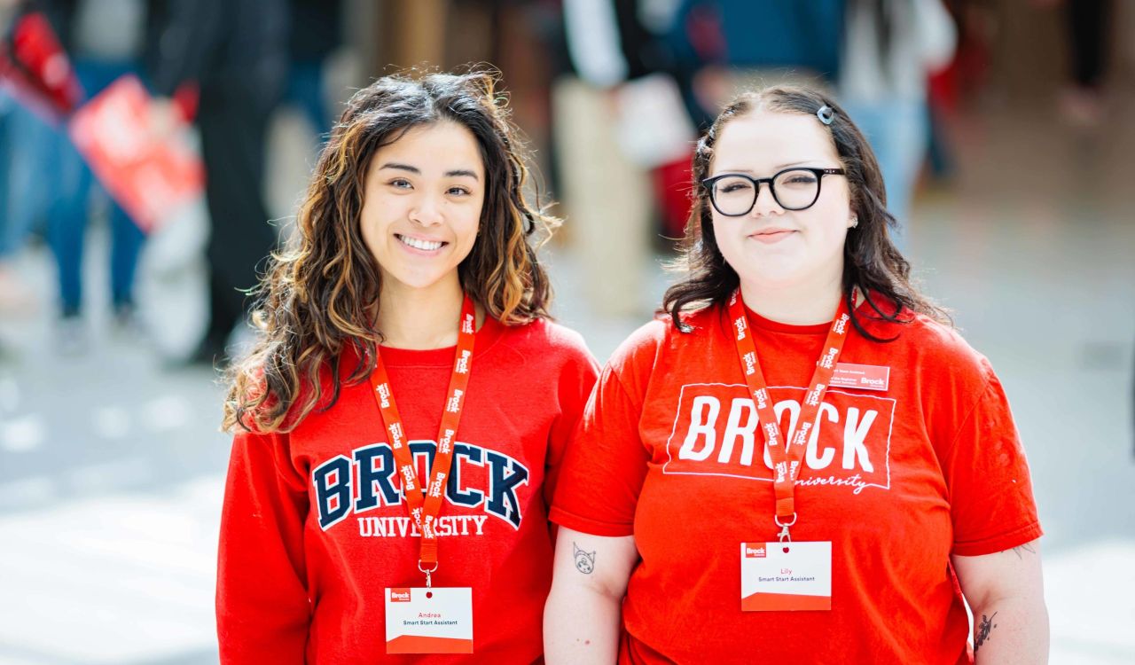 Two people in Brock University-branded shirts stand together in a busy room filled with people.