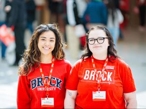 Two people in Brock University-branded shirts stand together in a busy room filled with people.