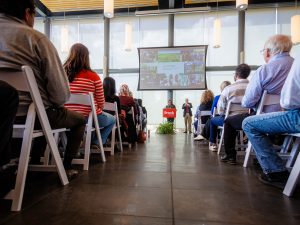 Audience members attend a Brock University event in a modern gathering space, where speakers present from a podium beside a large screen displaying presentation slides.