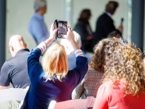 Audience members sit in rows of white chairs at an indoor event while one attendee holds a phone above their head to capture a QR code at the front of the room. The speaker and other people in the background are out of focus, with bright sunlight filling the space.