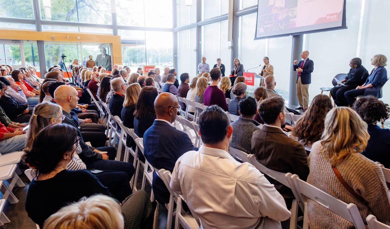 A large audience gathers in a sunlit atrium with floor-to-ceiling windows for a Brock University event. Speakers and panelists sit and stand at the front of the room while attendees face a large screen displaying presentation content.