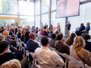 A large audience gathers in a sunlit atrium with floor-to-ceiling windows for a Brock University event. Speakers and panelists sit and stand at the front of the room while attendees face a large screen displaying presentation content.