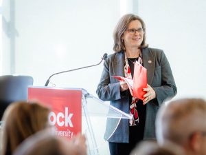 A woman holds a red folder in her hands while standing in front of a podium with Brock University branding and a crowd of people.
