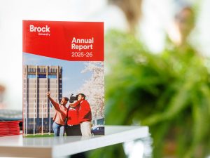 Brock University’s 2025-26 Annual Report stands upright on a table, with the cover showing students and the University mascot in front of Schmon Tower. Blurred greenery and people appear in the background, with additional copies of the report stacked beside it.
