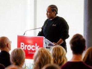 A woman stands at a Brock University branded podium speaking in front of a crowd.