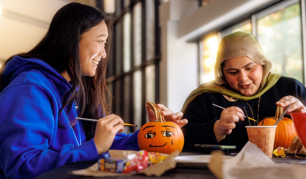 Two students paint pumpkins together at a table, decorating them with colourful designs while enjoying a fall activity indoors.
