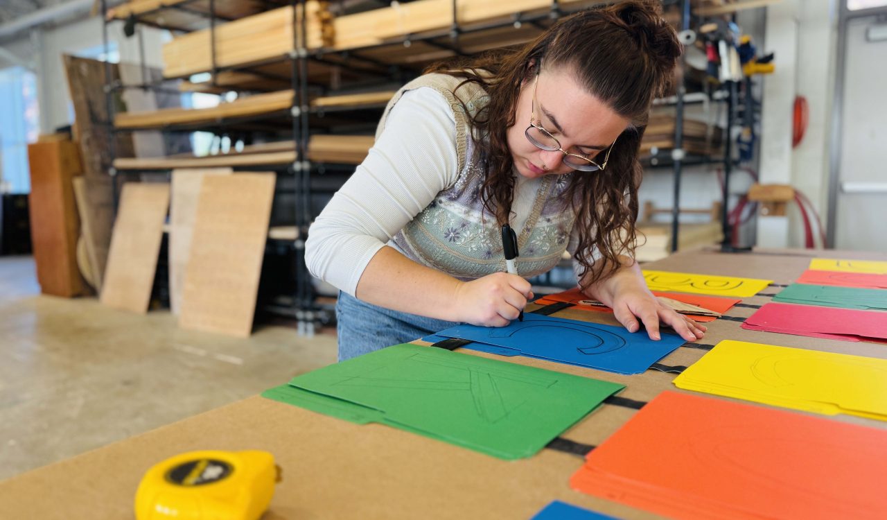 Em Schilling leans over a large drafting table and draws on colourful sheets of paper in Brock’s scene shop at the Marilyn I. Walker School.