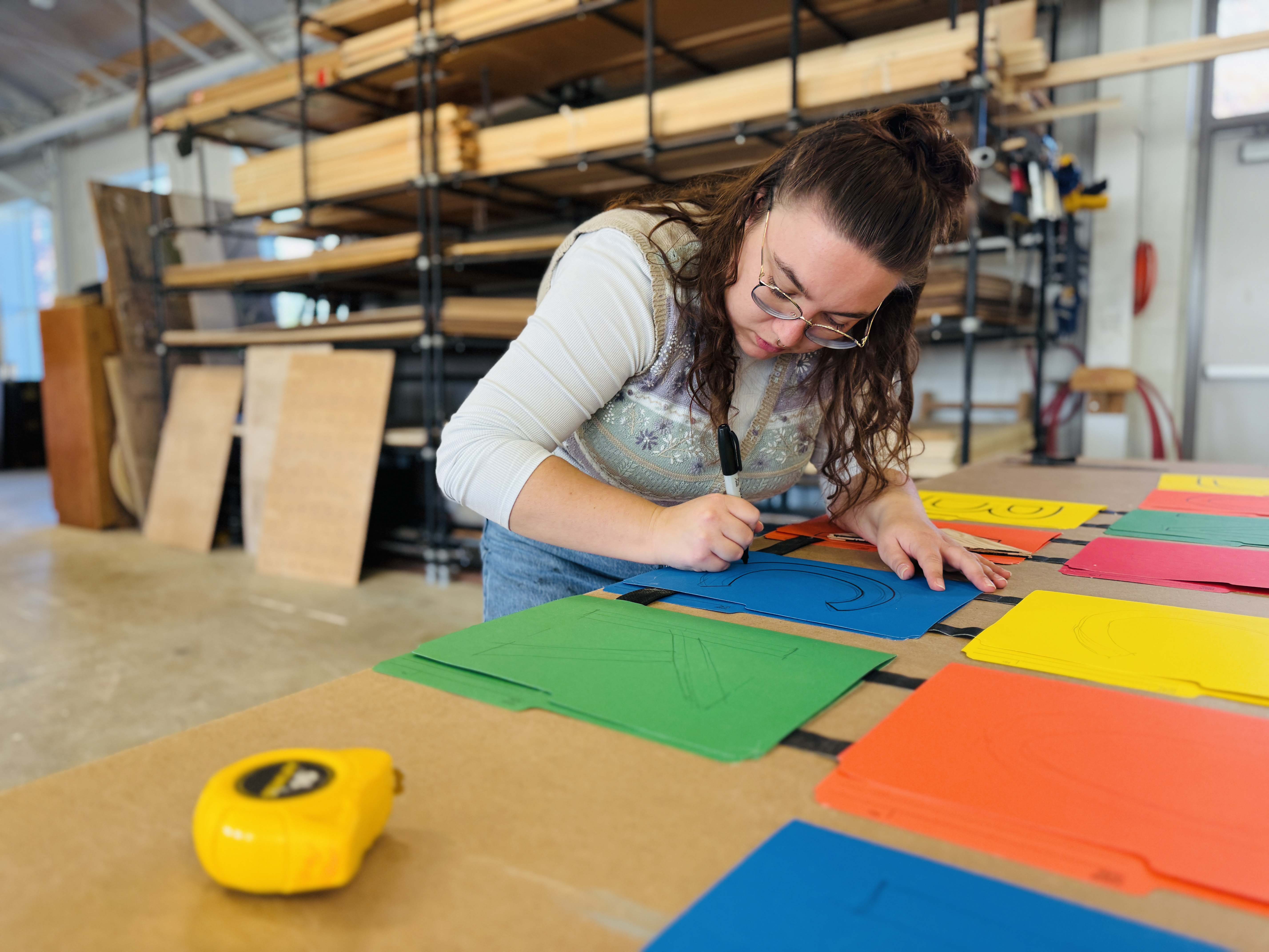 Em Schilling leans over a large drafting table and draws on colourful sheets of paper in Brock’s scene shop at the Marilyn I. Walker School.