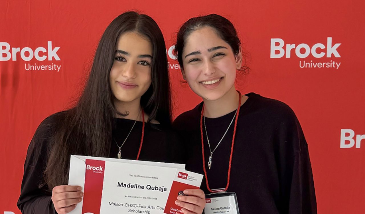 There are two women standing dressed in black tops and blue jeans against a red backdrop with the words “Brock University.”