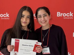 There are two women standing dressed in black tops and blue jeans against a red backdrop with the words “Brock University.”