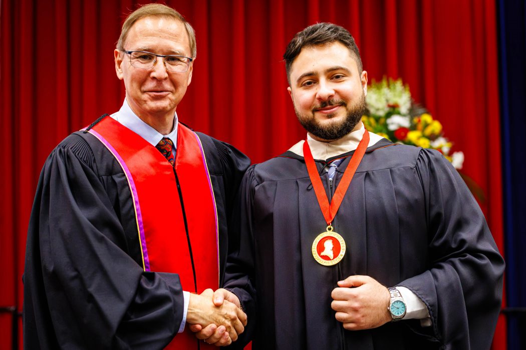 Two people in Convocation gowns stand together shaking hands. One is wearing a medal around his neck.