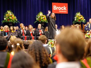 A graduate waves to the crowd from the stage while a seated procession looks on.