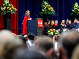 A woman stands at a podium speaking to a crowd at a graduation ceremony.