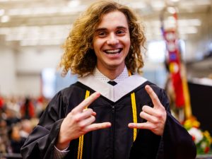 A man in a graduation gown lifts his index and ring fingers on both hands while smiling.