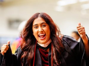 A woman in a Convocation gown gestures excitedly while smiling.
