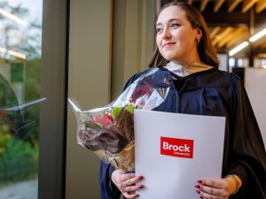 A woman in a graduation gown holds a bouquet of flowers and a Brock University-branded folder.